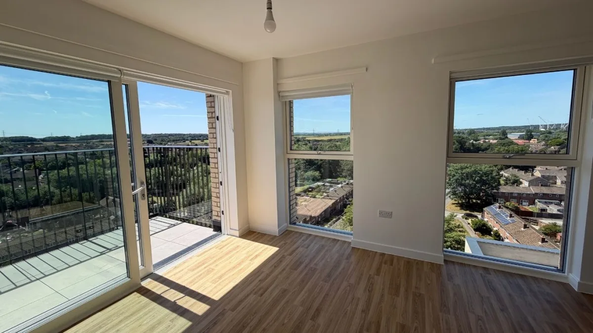 An empty new flat with wood-effect flooring, a glazed door on to a balcony and two large windows looking out over houses, trees and fields