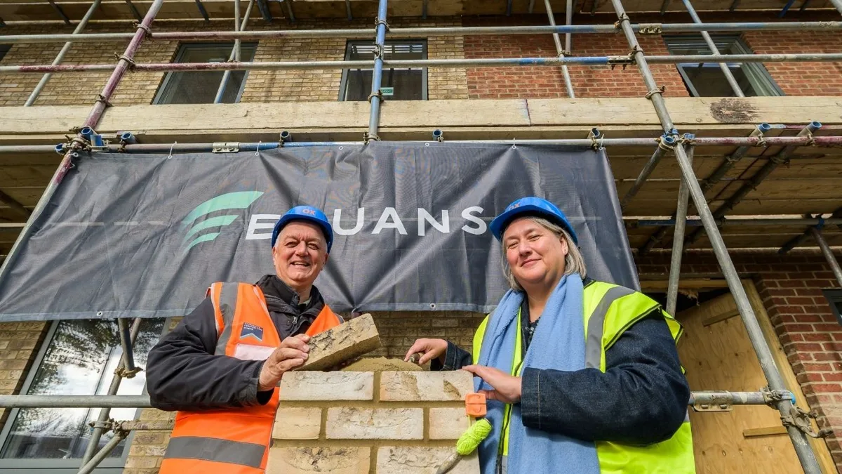 Two people in hard hats and high-visibility jackets lay a brick in front of scaffolding and an Equans banner at the Northfield Gate housing site in Littlemore.