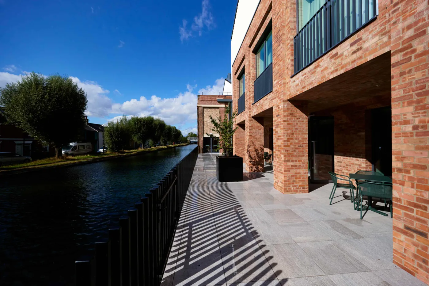 A paved riverside terrace beside the brick building, with tables and chairs and the river running alongside