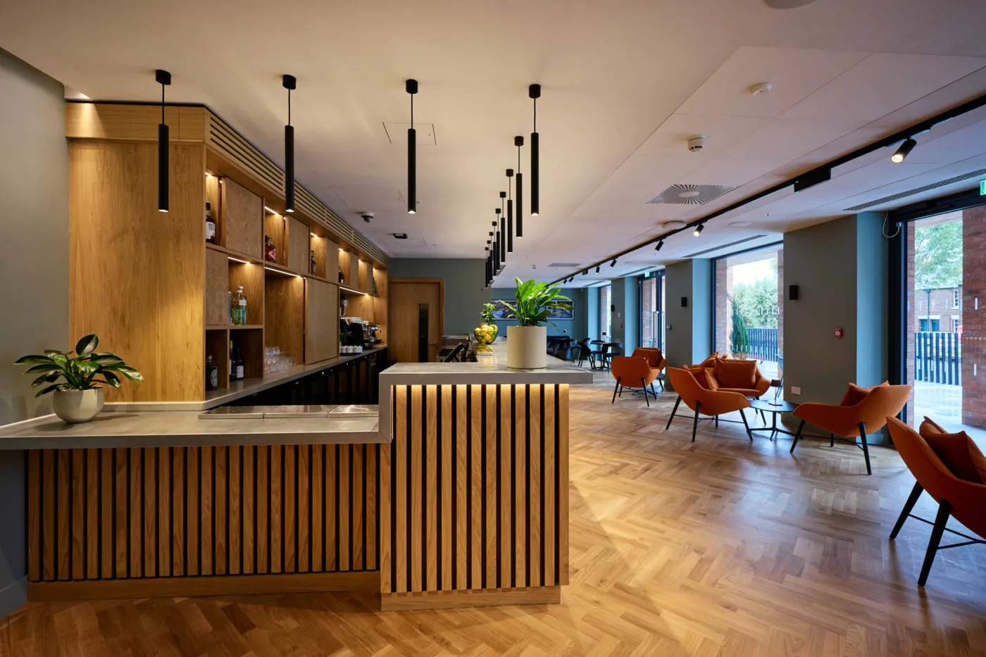 A clubroom bar with wooden panelling, pendant lights and orange chairs by tall windows
