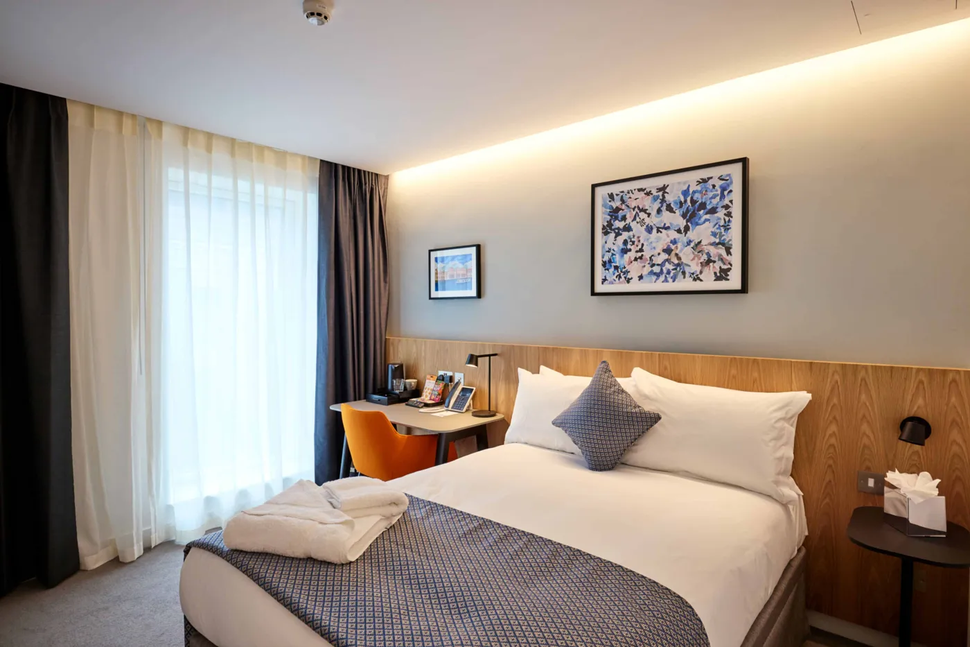 A bedroom with a double bed, a desk and chair, and a window with long curtains