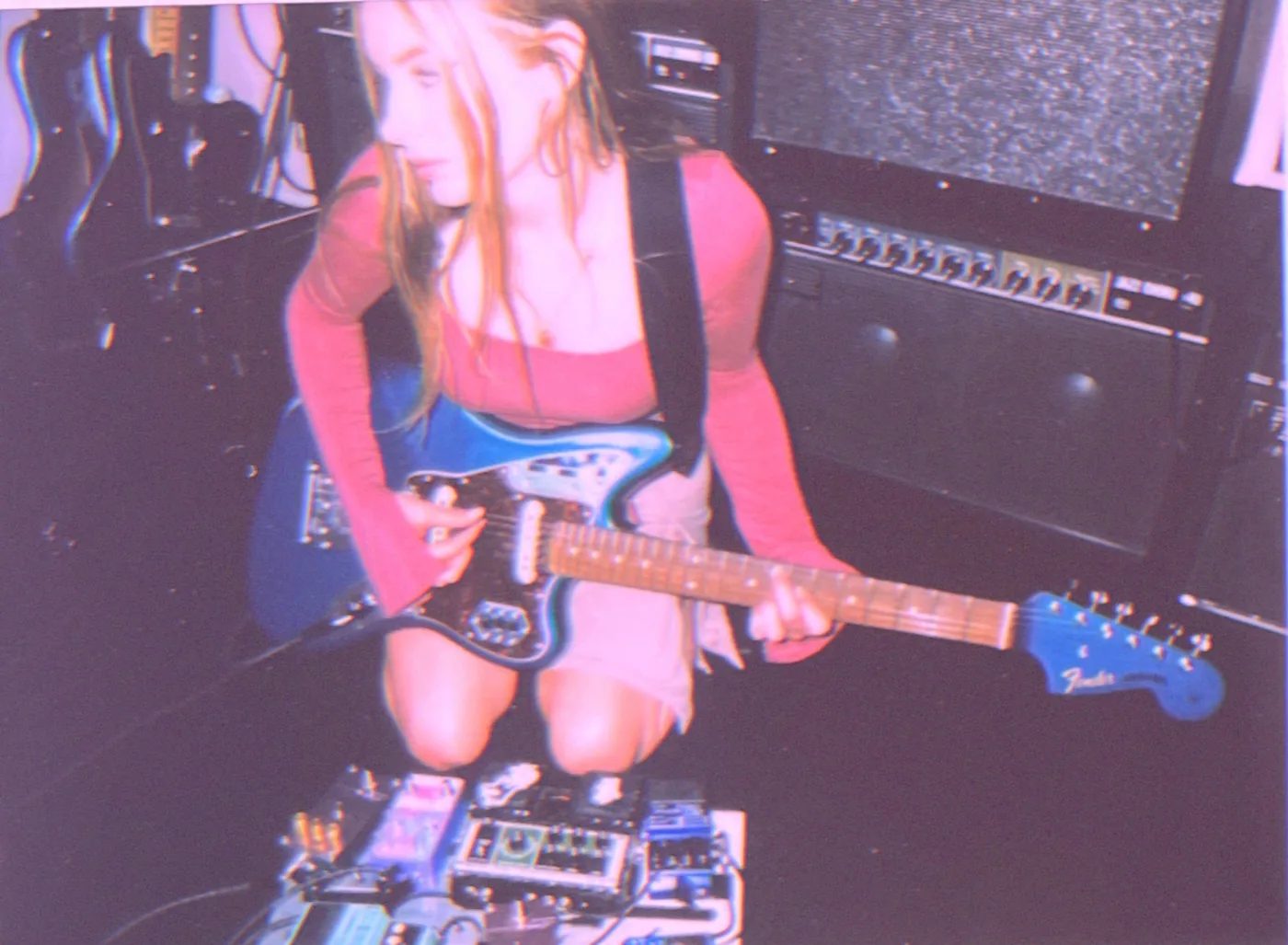 Paige of abstract in sound kneeling with a blue electric guitar beside a pedalboard of effects pedals, amplifiers behind her