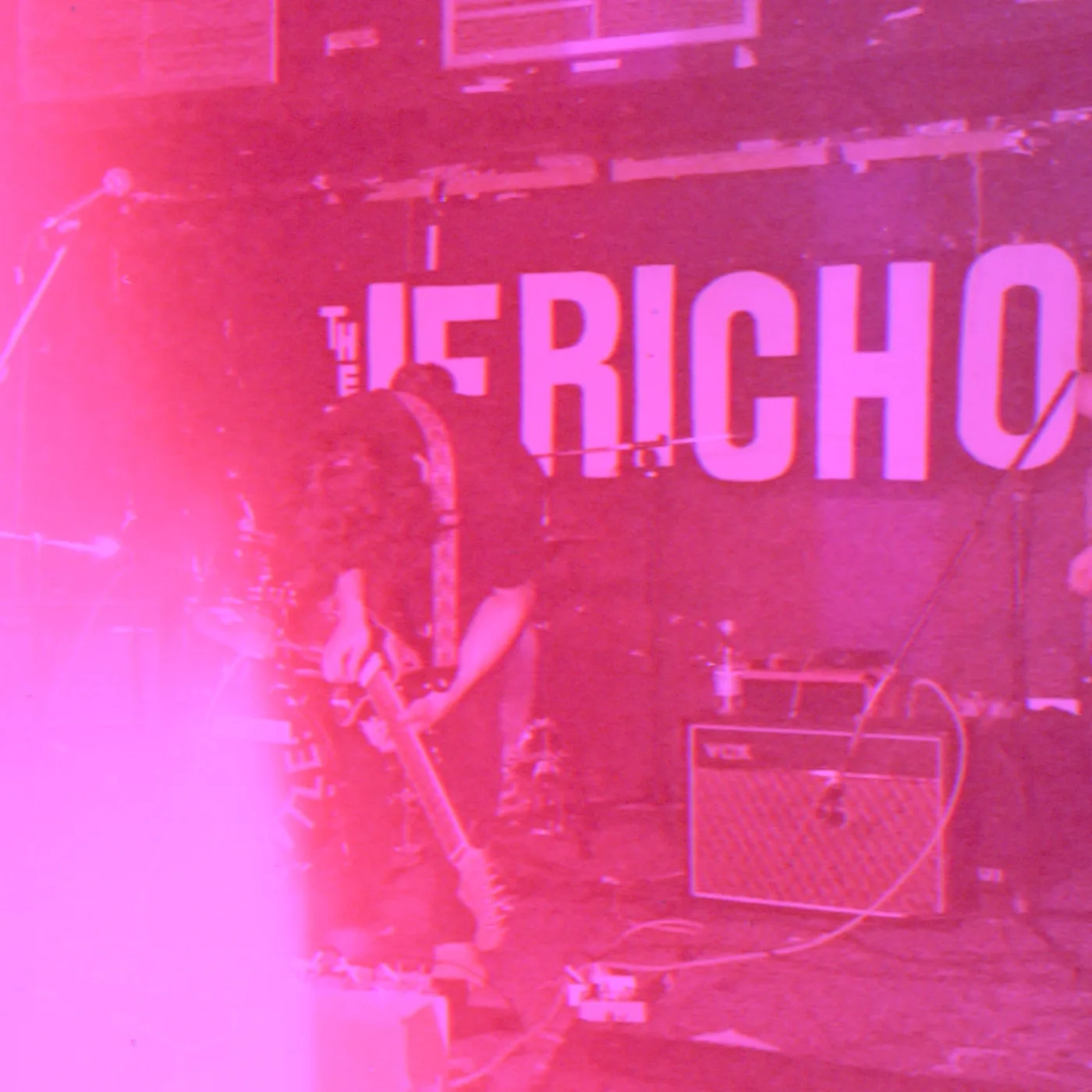 A guitarist playing on a dimly lit stage in front of a large sign reading The Jericho, with an amplifier beside him
